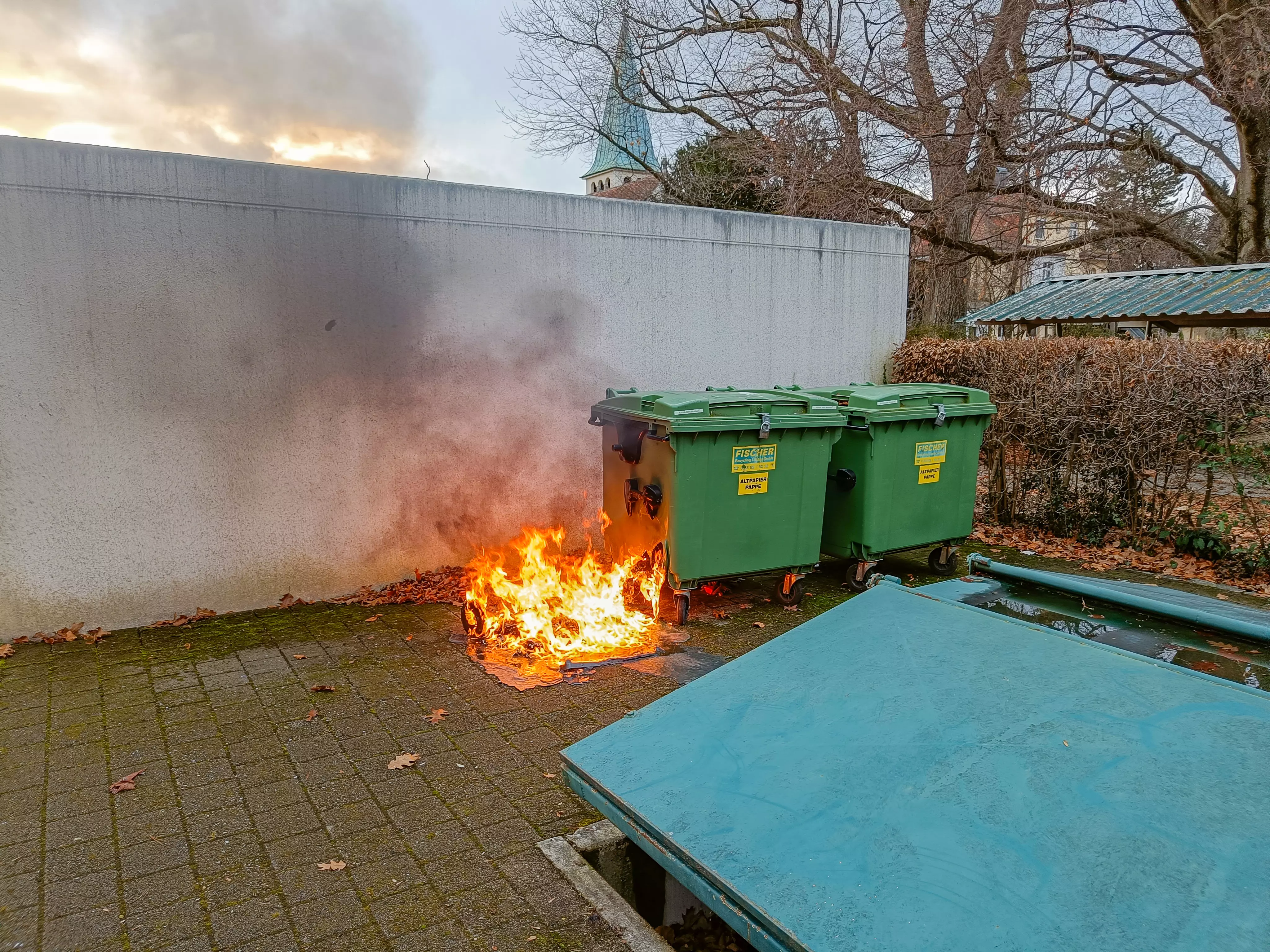 Ein Müllcontainer brennt neben zwei weiteren Müllcontainern. Direkt dahinter befindet sich eine Garage.