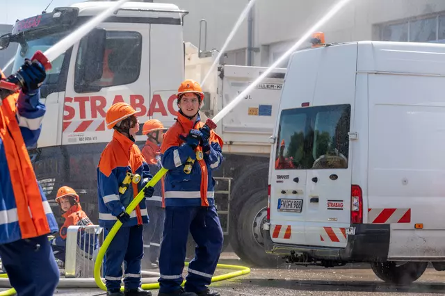 Drei Trupps der Jugendfeuerwehr löschen aus Strahlrohren den fiktiven Brand. Im Hintergrund Firmenfahrzeuge der Strabag GmbH.