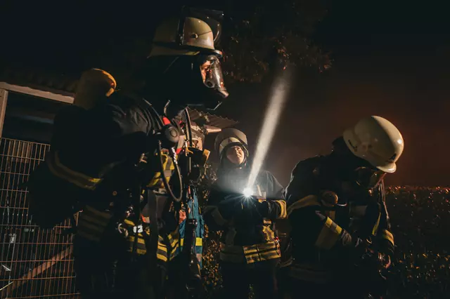 Ein Trupp unter Atemschutz bereitet sich für den Einsatz vor. Zwischen den beiden Kräften steht eine Feuerwehrfrau, die nach oben blickt. Dorthin führt auch der Lichtkegel ihrer Handlampe.