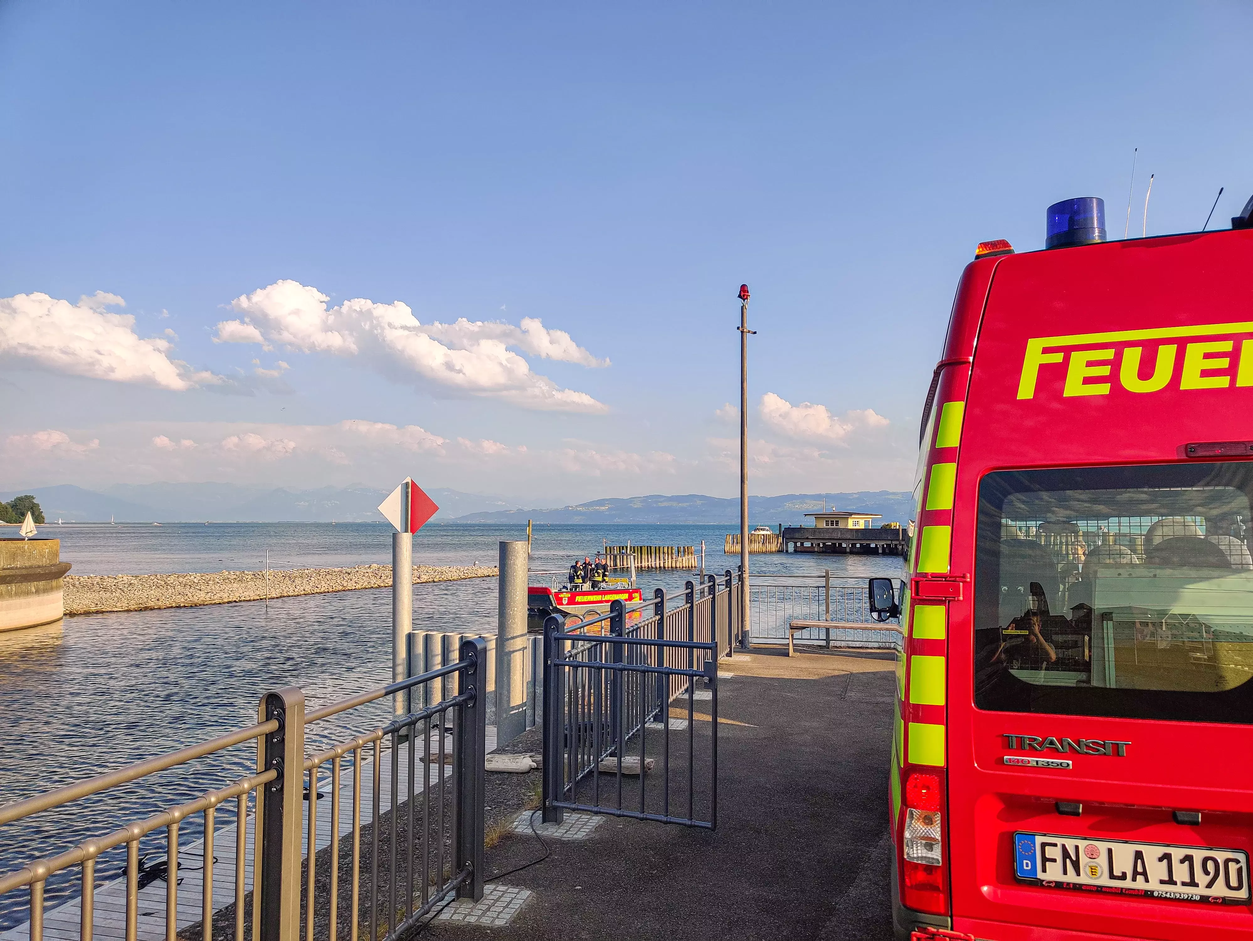 Rechts am Bildrand steht der MTW 1 der Feuerwehr Langenargen im Vordergrund, links davon ist die Hafeneinfahrt zu sehen, die gerade vom Mehrzweckboot befahren wird. Im Hintergrund der Bodensee bei strahlendem Sonnenschien.