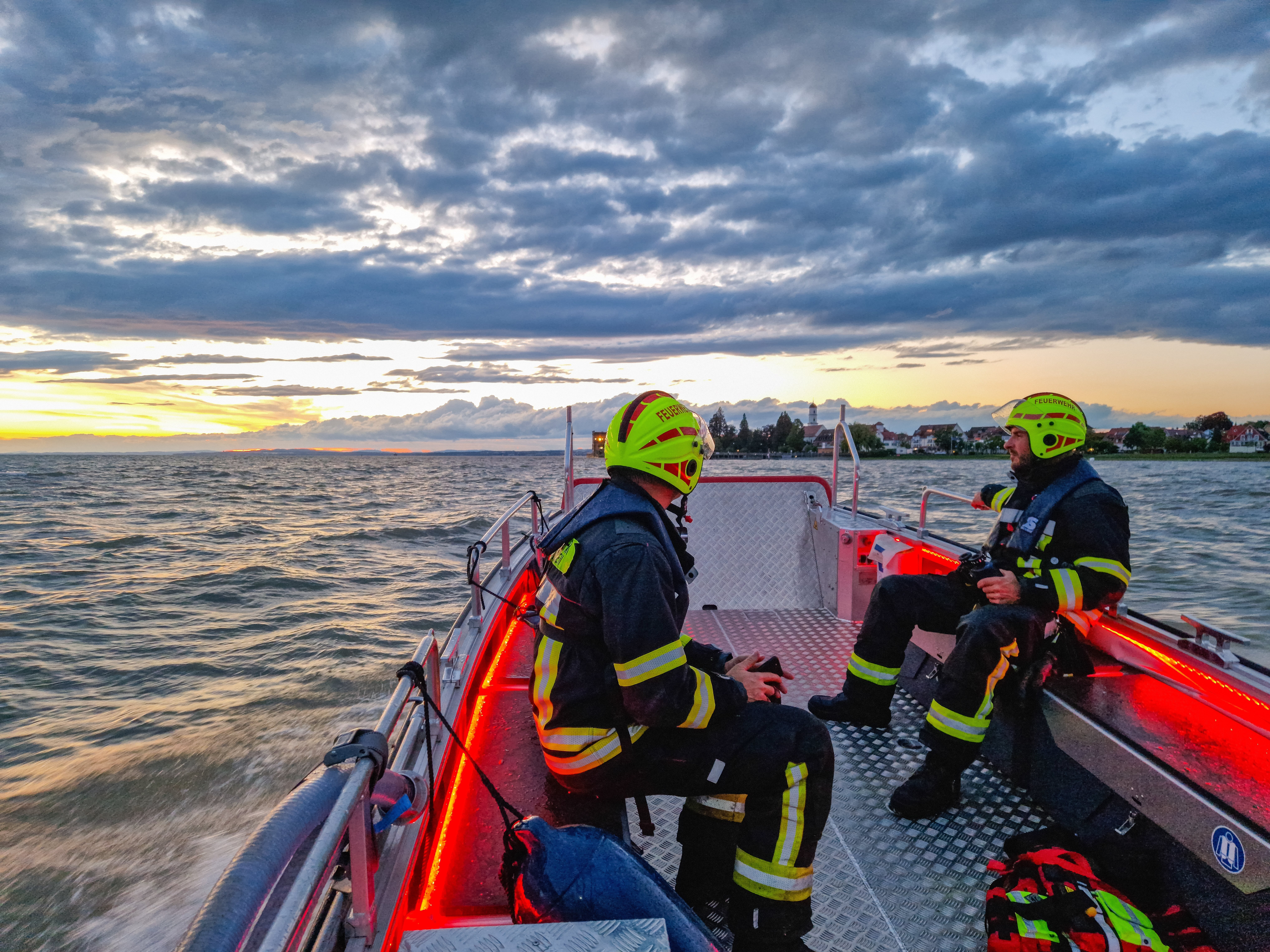 Das Mehrzweckboot ist auf der Rückfahrt von Kressbronn in Richtung Langenargen auf dem Bodensee. Im Vordergrund sind zwei Einsatzkräfte zu sehen, im Hintergrund das Bodenseeufer mit Schloss Montfort.