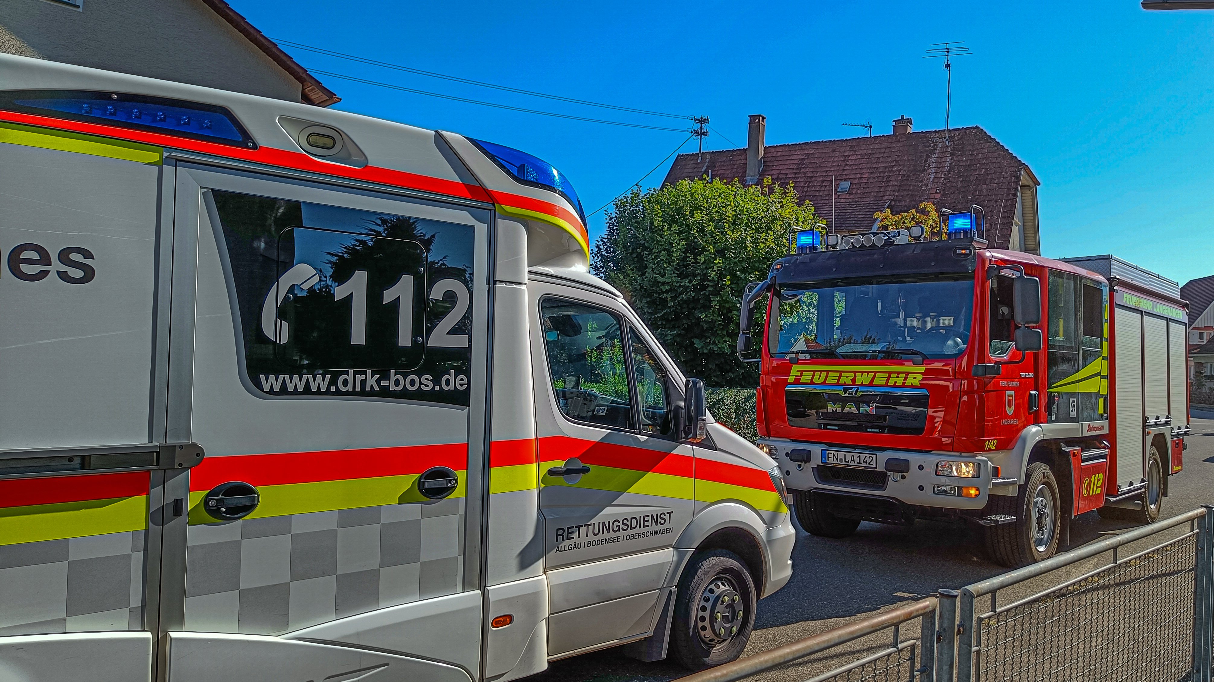 Rettungswagen und Löschfahrzeug stehen mit Blaulicht auf einer Straße. Blauer Himmel und Sonnenschein.