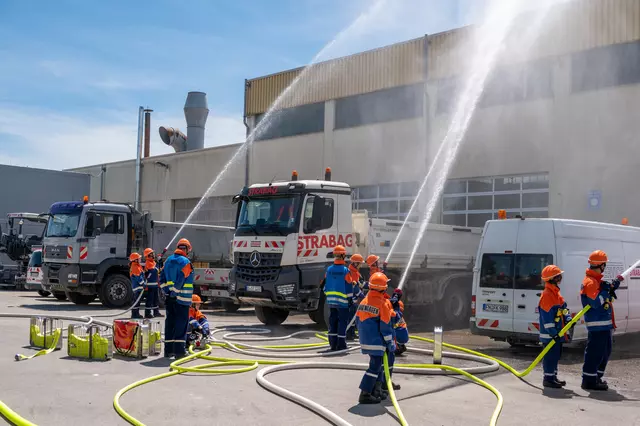 Mehrere Jugendfeuerwehrler löschen den fiktiven Brand einer Halle, die im Hintergrund zu sehen sind. Ebenfalls sind mehrere LKWs der Firma Strabag.