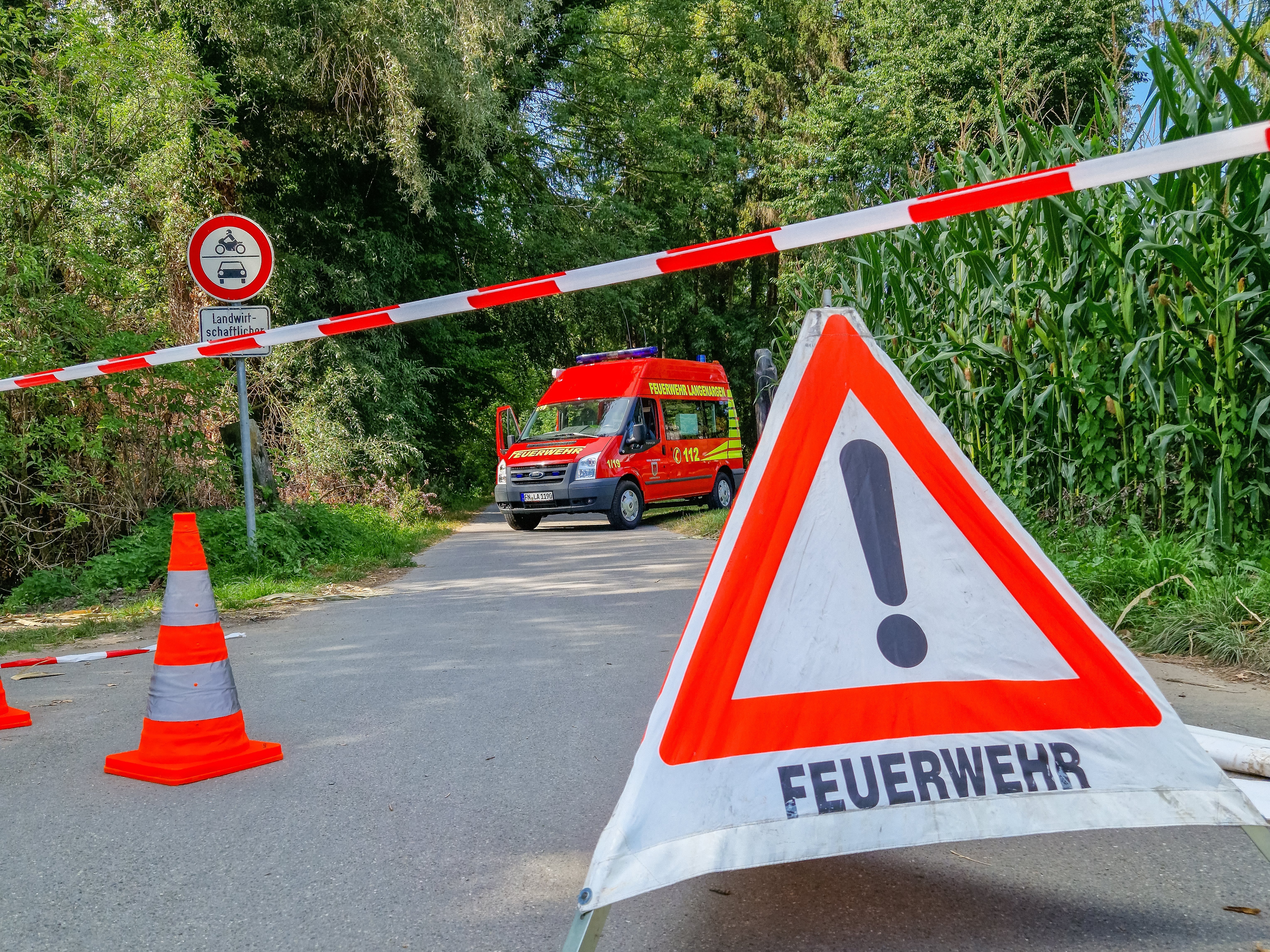 Ein Faltdreieck mit der Aufschrift Feuerwehr und eine Pylone sind im Vordergrund zu sehen. Der Weg ist mit Flatterband abgesperrt. Im Hintergrund steht der MTW quer auf dem Weg.