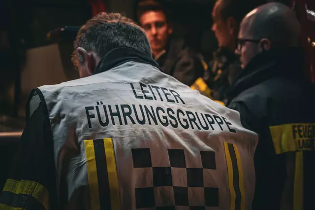 Rückenansicht der Funktionsweste "Leiter Führungsgruppe". Im Hintergrund sind mehrere Einsatzkräfte der Gruppe Information und Kommunikation zu sehen.