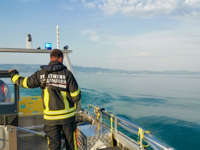 Im Vordergrund das MZB 1/79, darauf ein Feuerwehrmann mit Jacke. Im Hintergrund ist der Rettungshubschrauber über dem Bodensee zu sehen.