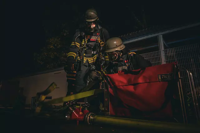 Zwei Feuerwehrmänner unter Atemschutz bereiten ihren Einsatz vor. Sie schließen einen Schlauchtragekorb an den Verteiler an. 