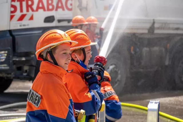 Im Vordergrund sind zwei Jugendfeuerwehrler mit Löschen beschäftigt. Im Hintergrund sieht man einen weiteren Trupp, sowie einen LKW der Firma Strabag.