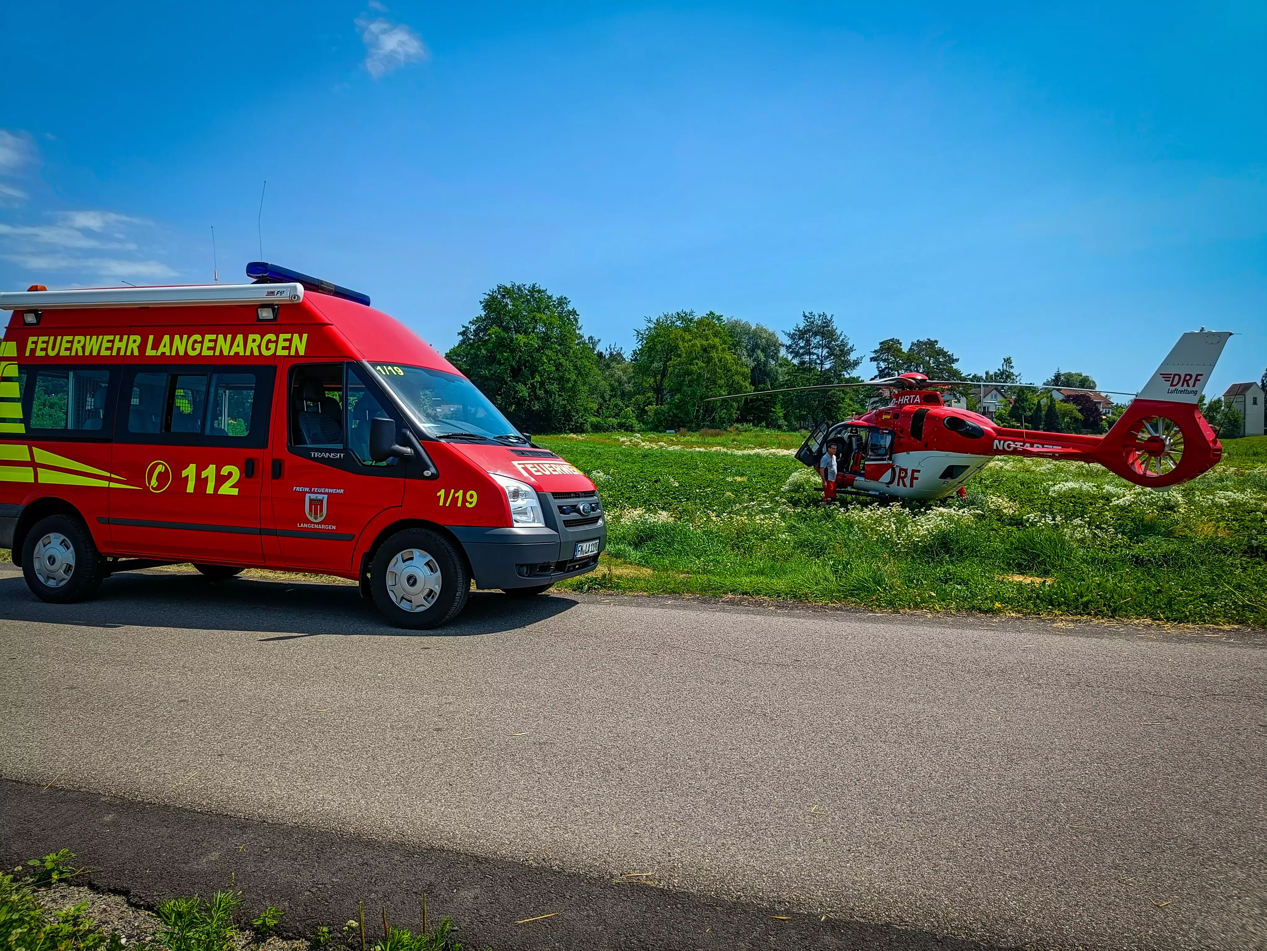 Der MTW 1 steht im Vordergrund, im Hintergrund ist der Rettungshubschrauber Christoph 45 aus Friedrichshafen zu sehen. Die Sonne scheint, der Himmel ist blau.