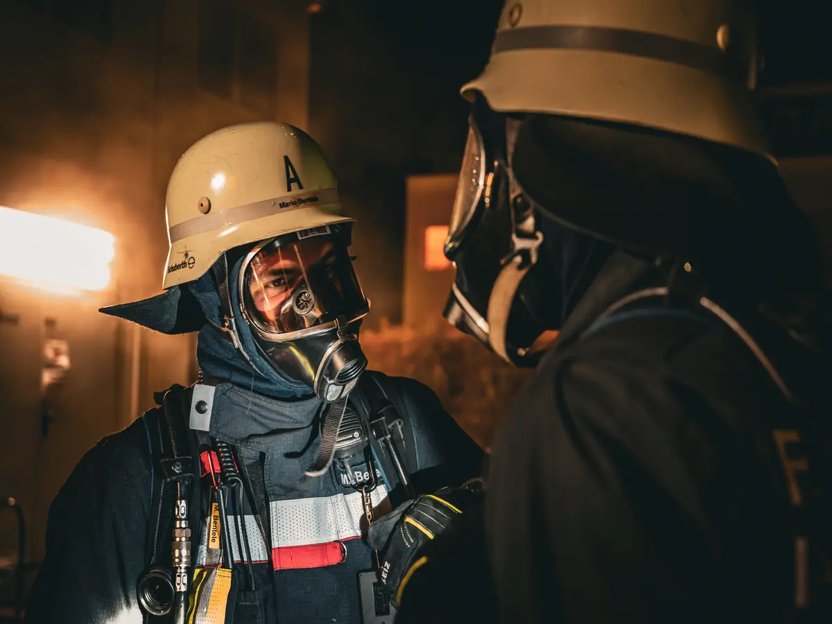 Zwei Feuerwehrmänner unter Atemschutz stehen sich gegenüber. Im Hintergrund ist eine Rauchentwicklung erkennbar.