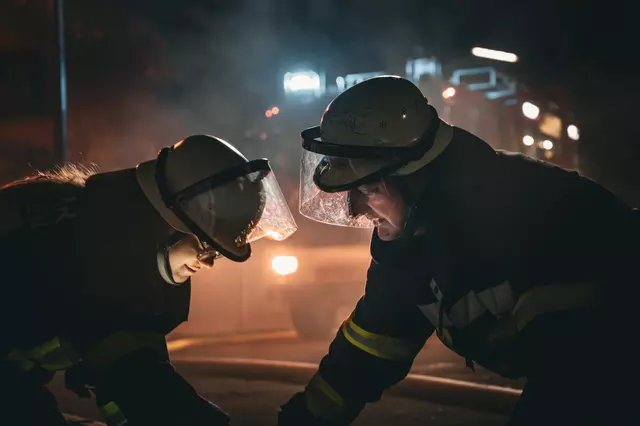 Zwei Feuerwehrangehörige sind im Vordergrund dabei, eine Steckleiter einsatzklar zu machen. Im Hintergrund das Löschfahrzeug 1/44 mit Blaulicht.