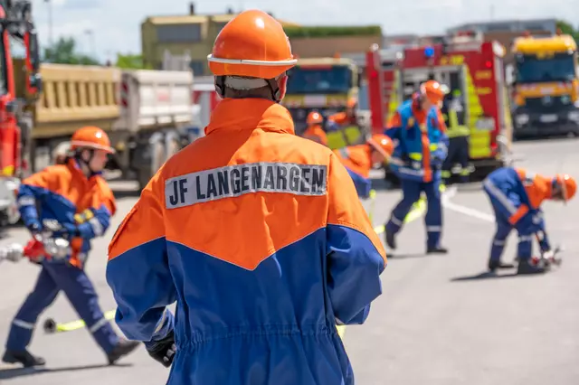 Ein Jugendfeuerwehrmitglied ist von hinten zu sehen. Auf der Jacke ist die Aufschrift JF Langenargen zu sehen. Im Hintergrund bereiten mehrere Jugendliche den Löschangriff vor.